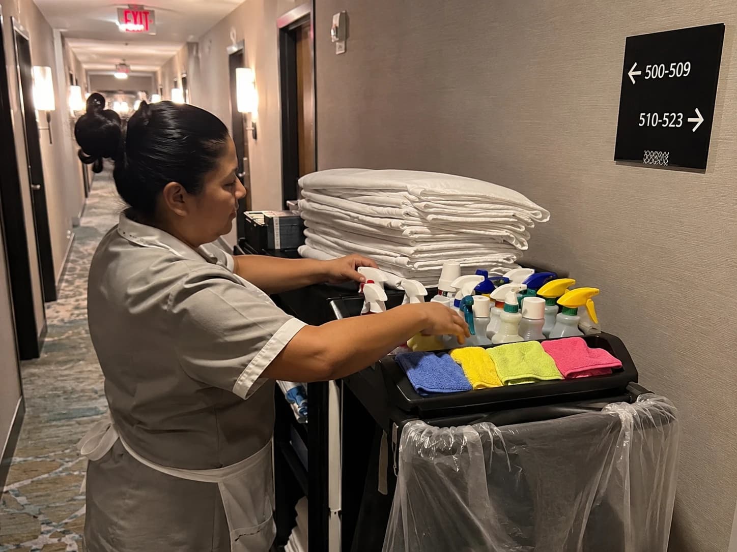 Femme de chambre préparant un chariot de nettoyage dans un couloir d'hôtel, avec draps pliés et produits d'entretien organisés par code couleur