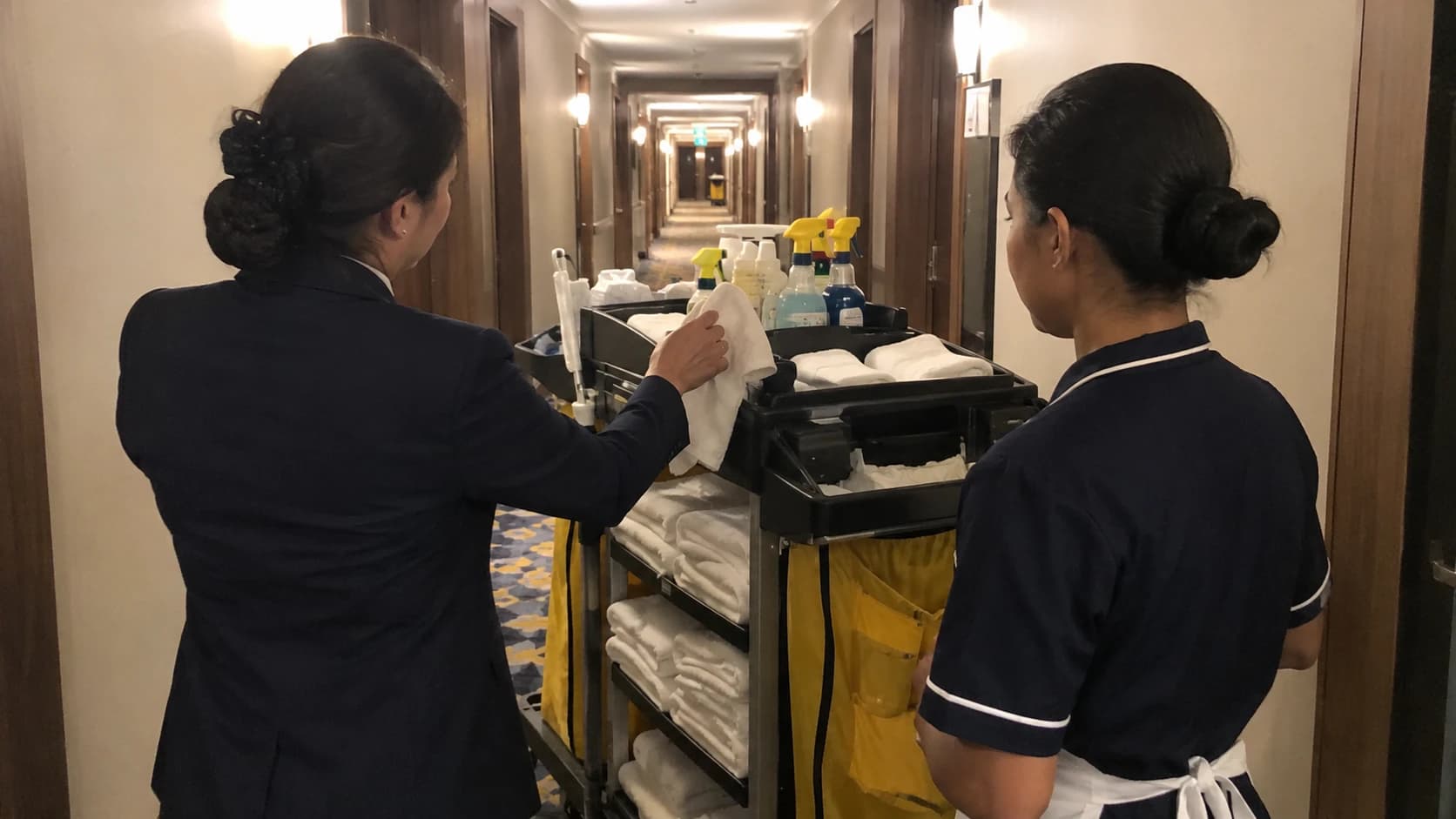 Gouvernante d'hôtel formant une nouvelle femme de chambre devant un chariot de nettoyage dans un couloir hôtelier professionnel