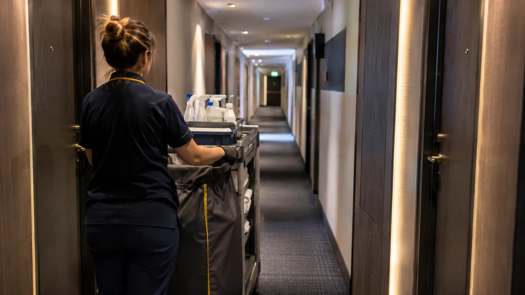 Femme de chambre de dos préparant un chariot de nettoyage dans le couloir d'un hôtel d'affaires lillois, touches de jaune et de bleu nuit