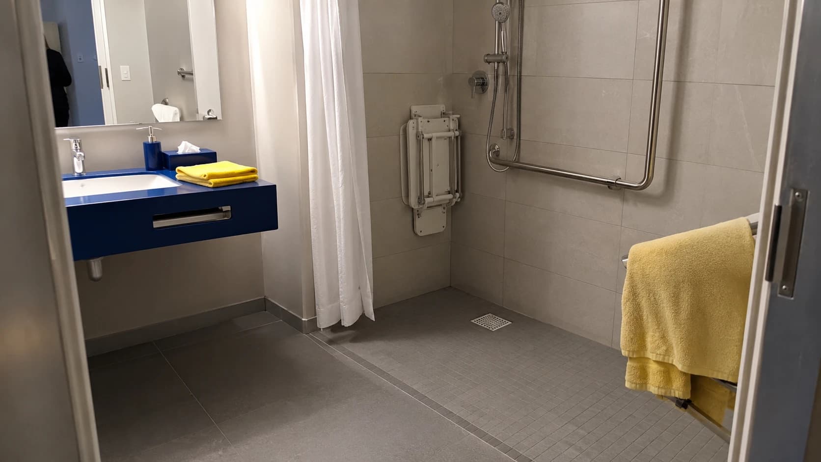 Salle de bains PMR d'hôtel avec barre d'appui et siège de douche relevable impeccables, serviette jaune pliée posée à portée de main