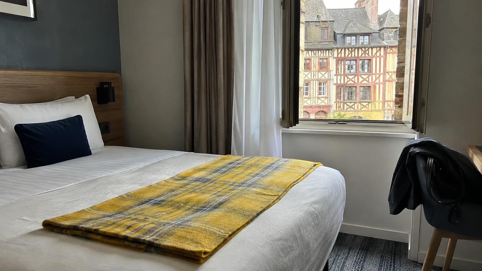 Chambre d'hôtel rennaise fraîchement préparée avec vue sur les façades à colombages du centre historique de Rennes, plaid jaune plié sur le lit