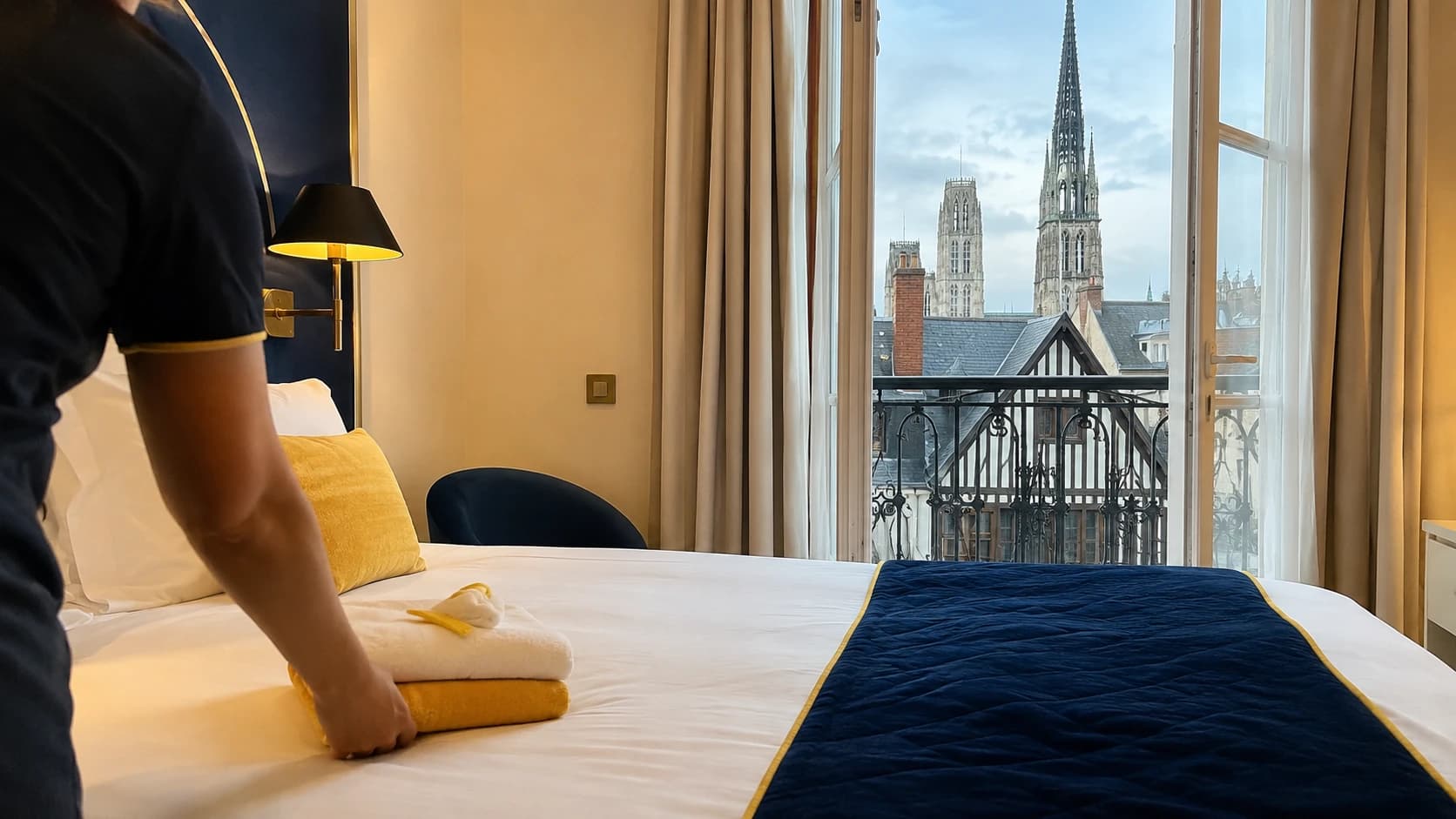 Chambre d'hôtel rouennaise fraîchement préparée avec vue sur les toits du centre historique et la cathédrale, serviette jaune pliée sur le lit, ambiance tourisme culturel normand