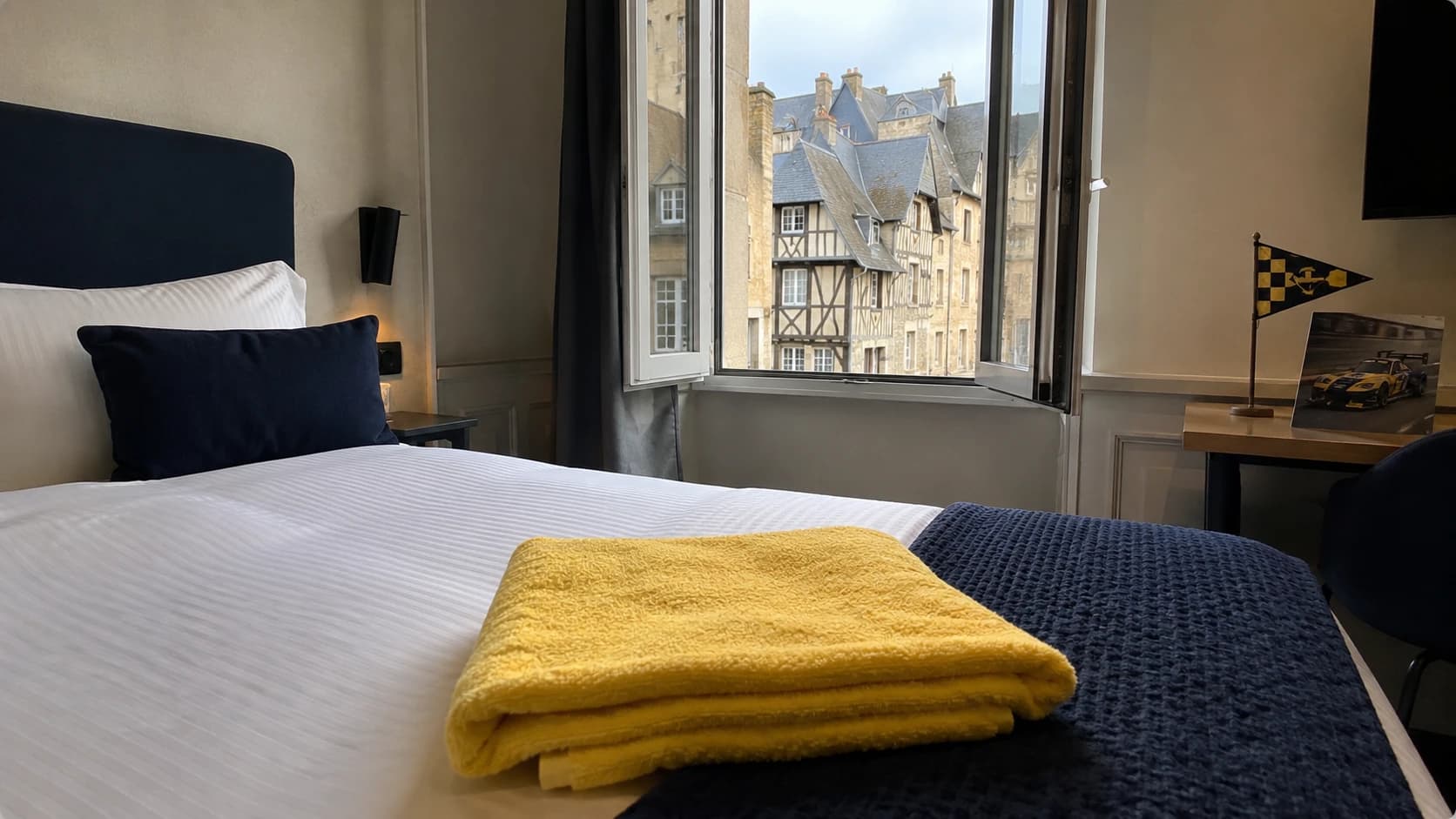 Chambre d'hôtel mancelle fraîchement préparée avec serviette jaune pliée sur le lit et vue sur les maisons à pans de bois de la Cité Plantagenêt, ambiance week-end des 24 Heures du