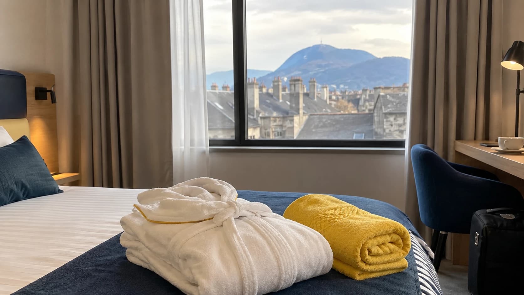 Chambre d'hôtel clermontoise fraîchement préparée avec vue sur le puy de Dôme, peignoir blanc et serviette jaune pliés sur le lit, ambiance de station thermale