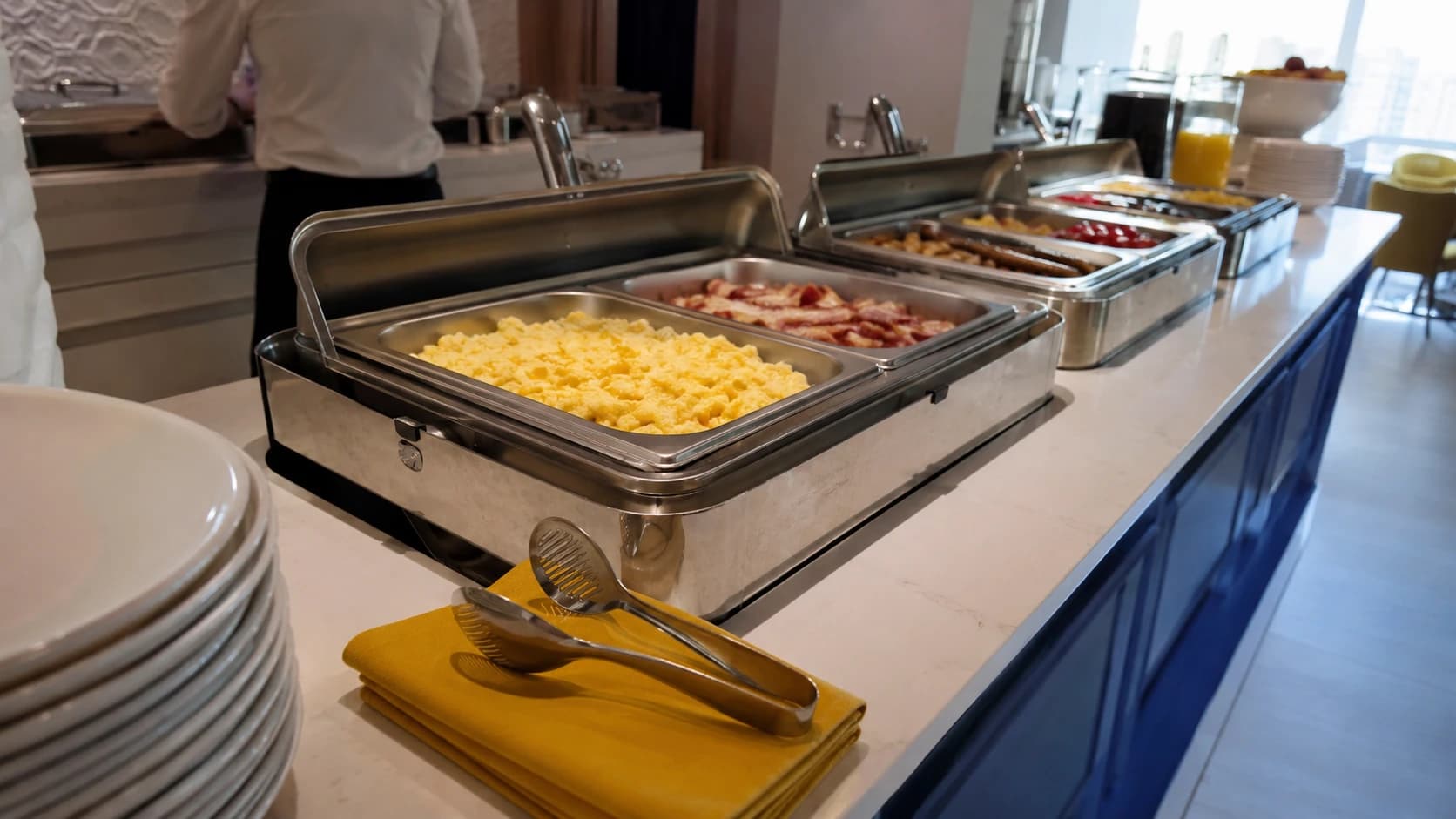 Buffet de petit-déjeuner d'hôtel fraîchement dressé avec bacs inox propres et pince de service posée sur un linge jaune plié